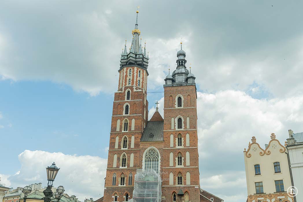 La Basílica de Santa María es uno de los monumentos más emblemáticos de Cracovia y una visita imprescindible en la Plaza del Mercado. Construida entre los siglos XIII y XIV sobre los restos de un templo anterior, destaca por su impresionante arquitectura gótica y por sus dos torres de diferente altura, una de las imágenes más reconocibles de la ciudad. En su interior alberga el espectacular retablo mayor tallado por Veit Stoss, considerado una de las obras maestras del arte gótico europeo. Cada hora, desde la torre más alta, un trompetista interpreta el Hejnał Mariacki, una melodía que se interrumpe bruscamente en recuerdo del centinela que, según la tradición, fue alcanzado por una flecha mientras alertaba a los ciudadanos de una invasión mongola en el siglo XIII. Esta emotiva costumbre, mantenida desde hace siglos, convierte a la basílica en un símbolo de la historia, la cultura y la identidad de Cracovia.